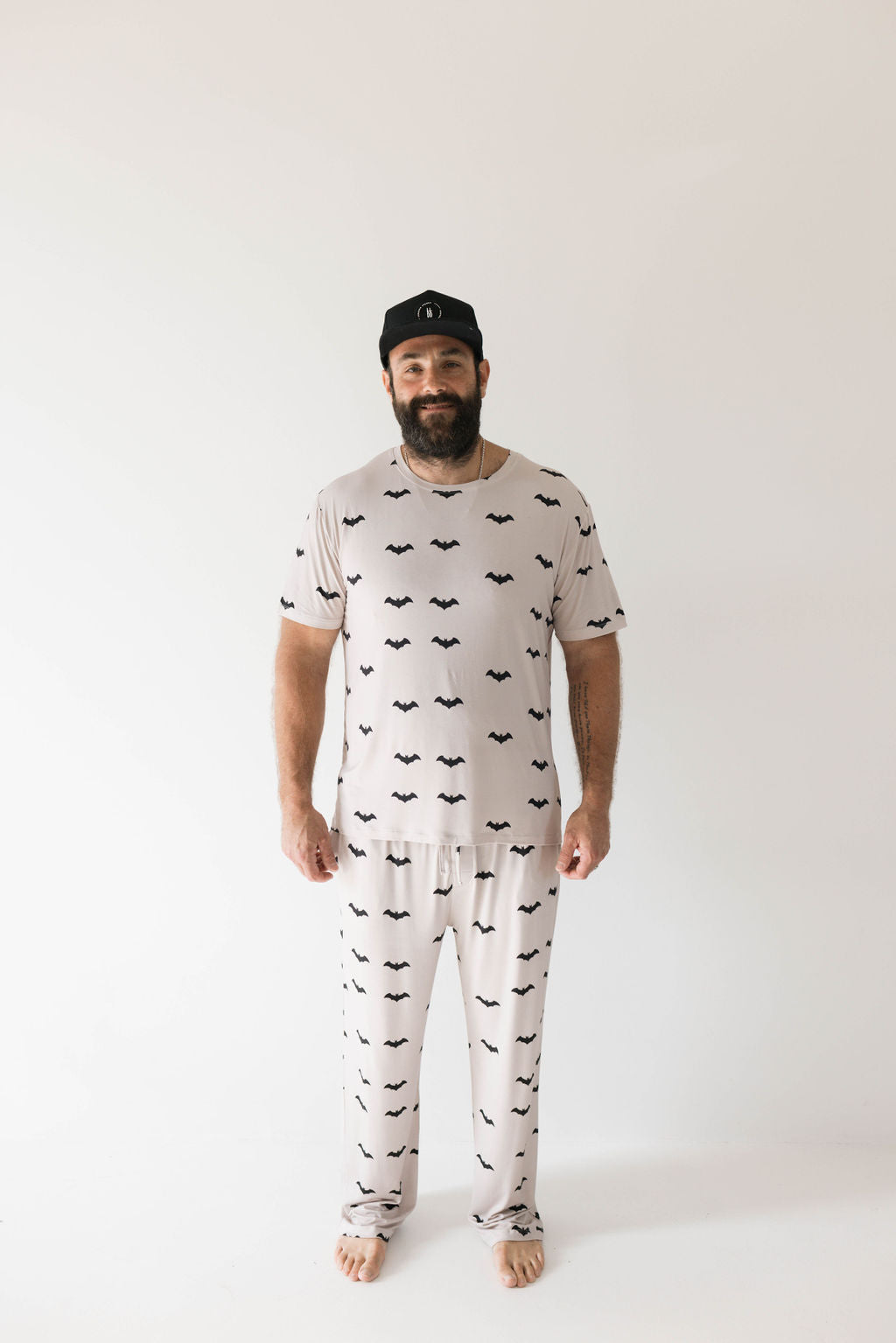 A man with a beard wearing a black cap stands against a plain white background. He is dressed in forever french baby's ultra-soft Adult Bamboo Short Sleeve Pajamas from their "It's Bats!" collection, featuring a pattern of small black bats. The hypo-allergenic clothing gives him a relaxed expression as he rests his hands by his sides.