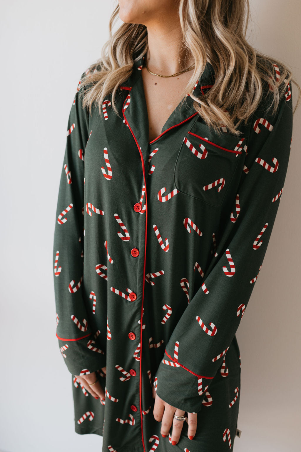 A woman stands wearing a Candy Cane Lane Women's Bamboo Sleeping Dress by forever french baby, featuring a dark green top adorned with red candy cane patterns and red piping. Her long, wavy hair gracefully frames her face. A ring glimmers on her hand, adding a touch of style. The light-colored background enhances the eco-friendly bamboo fabric of her attire, similar to a soothing night gown.