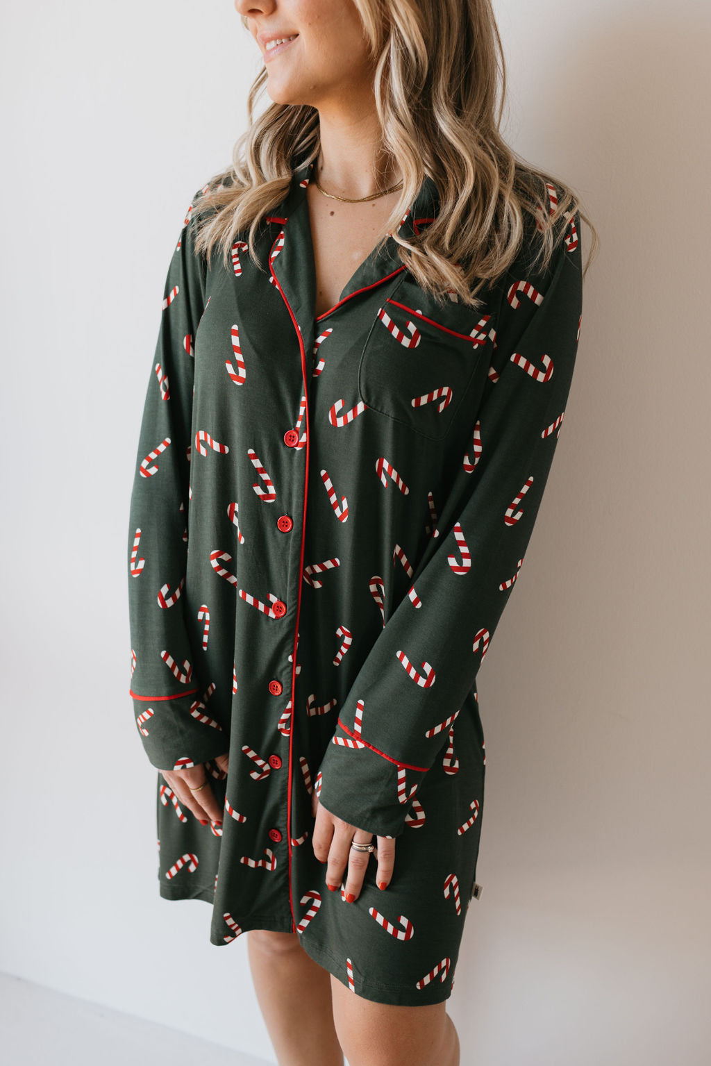 A woman models the forever french baby Women's Bamboo Sleeping Dress from the Candy Cane Lane collection. This long-sleeve, dark green nightshirt is adorned with festive candy cane patterns and highlighted by red piping and buttons. Made from eco-friendly bamboo fabric, it boasts hypo-allergenic properties. She stands against a white wall, her face angled slightly away from the camera.