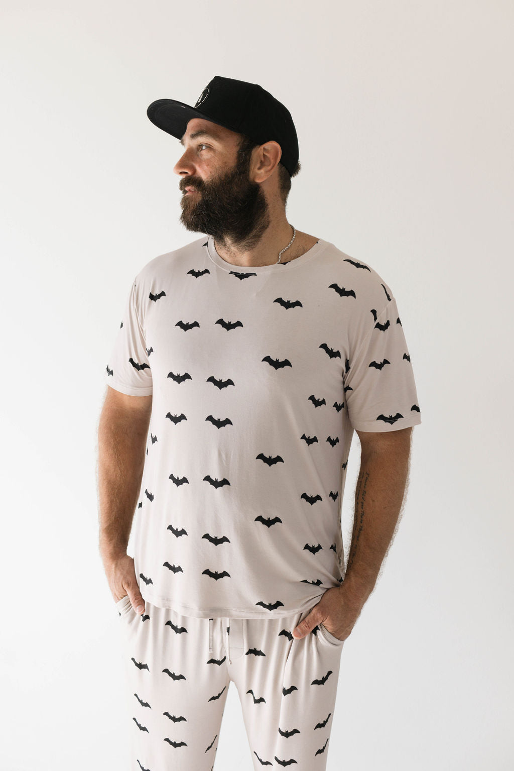 A bearded man stands against a plain background, looking to his right with his hands in his pockets. He is wearing a matching light-colored t-shirt and pants set adorned with black bat silhouettes made from ultra-soft bamboo fabric, known as the Adult Bamboo Short Sleeve Pajamas | It's Bats! by forever french baby. He also wears a black cap.