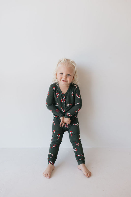 A smiling toddler with curly hair stands against a white wall, wearing the Bamboo Two Piece Pajamas | Candy Cane Lane from forever french baby. The hypo-allergenic material ensures comfort for sensitive skin, making every giggle even more delightful.