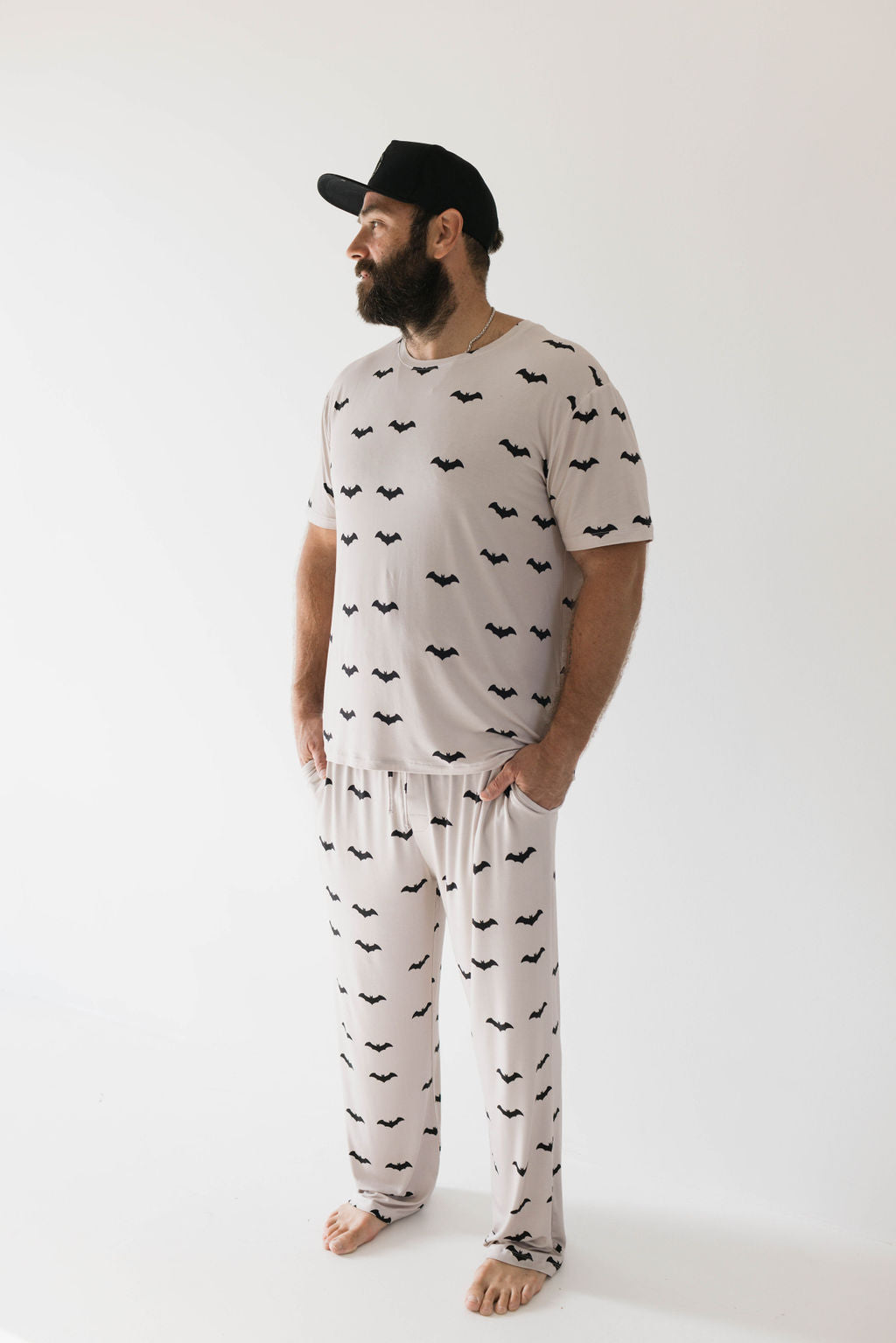 A man with a beard stands against a plain white background, wearing the ultra-soft "It’s Bats!" short sleeve pajama set from forever french baby, crafted from bamboo fabric and adorned with small black bat silhouettes. He is looking to the side and also wearing a black baseball cap.