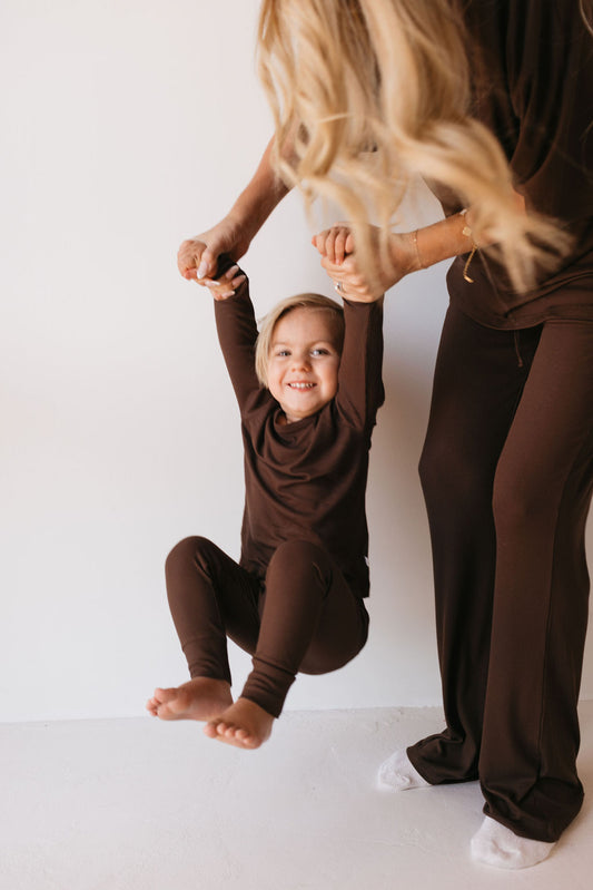 A smiling child, dressed in the Bamboo Two Piece Lounge Set in Coffee Bean by forever french baby, hangs by their arms, held by an adult with long blonde hair who is also wearing a matching brown outfit. They are indoors against a plain white background.