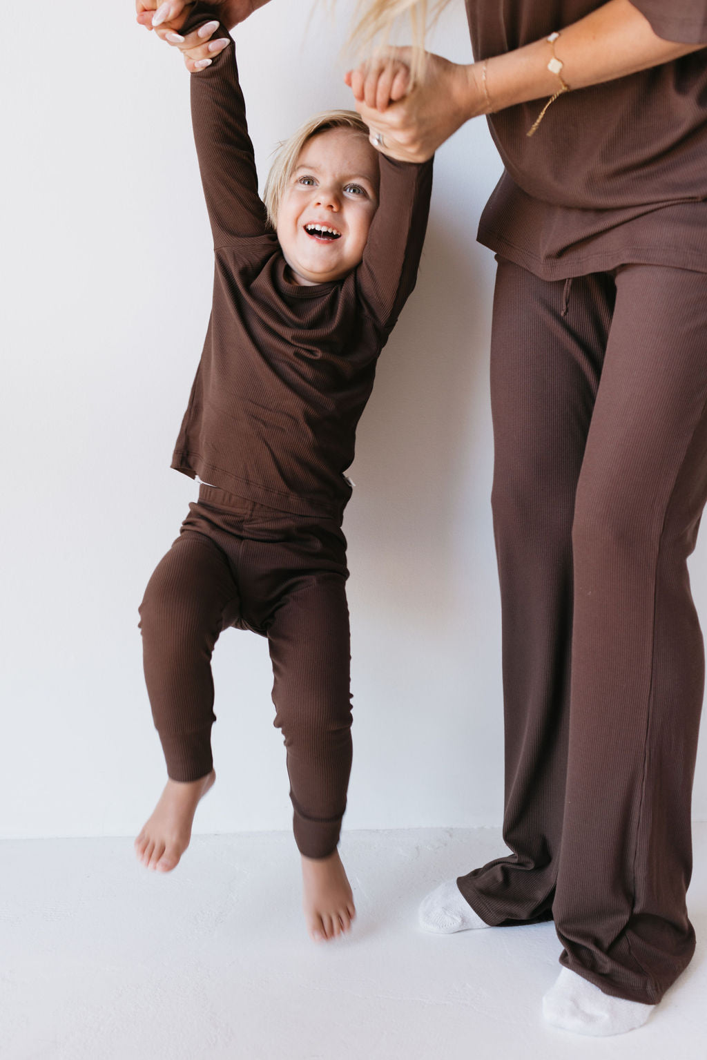 A joyful child dressed in the "Bamboo Two Piece Lounge Set | Coffee Bean" by forever french baby is being lifted playfully by a person wearing matching brown pants. The child's hands are held, and both appear to be having fun against a plain white background, enjoying the comfort of breathable fabric.