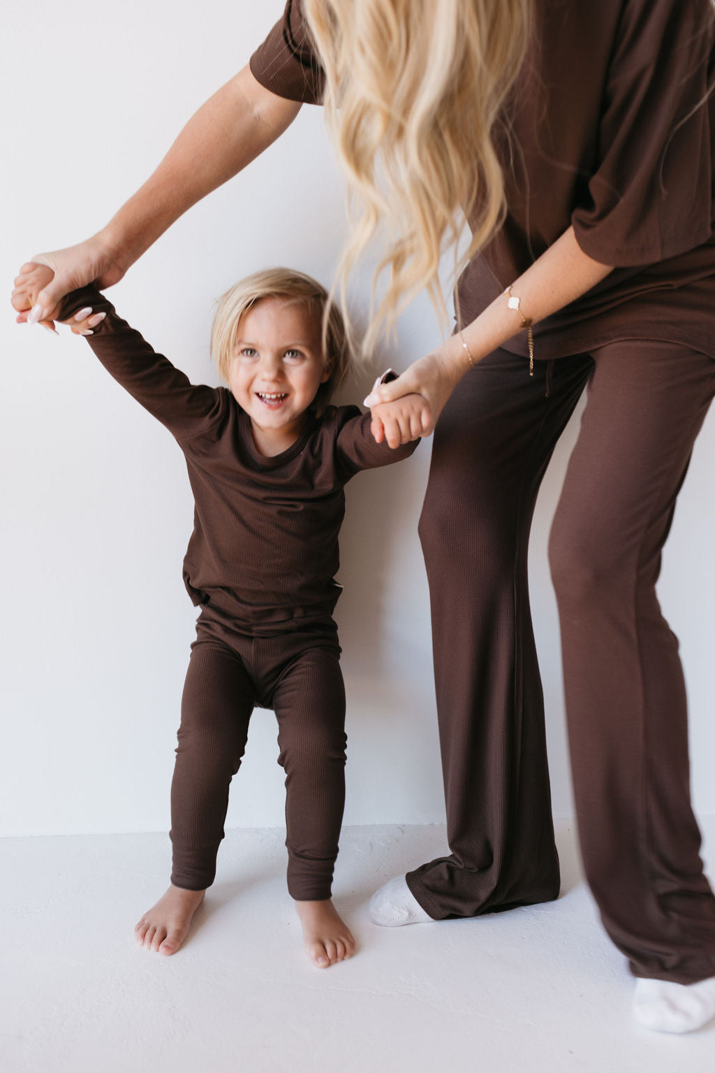 A young child with blonde hair, dressed in the Bamboo Two Piece Lounge Set in Coffee Bean by forever french baby, smiles happily while holding hands with an adult. The adult, also wearing a matching brown outfit, stands partially visible, helping the child keep balance. They stand on a light-colored floor.