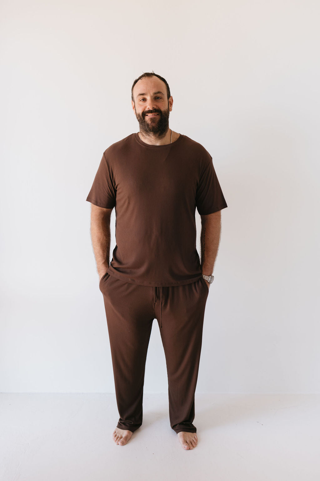 A person with a beard stands barefoot against a plain white background, showcasing the Men's Bamboo Short Sleeve Pajamas in Coffee Bean by forever french baby. The soft bamboo fabric of the matching chocolate brown T-shirt and pants complements their subtle, relaxed smile as they casually tuck their hands into their pockets.