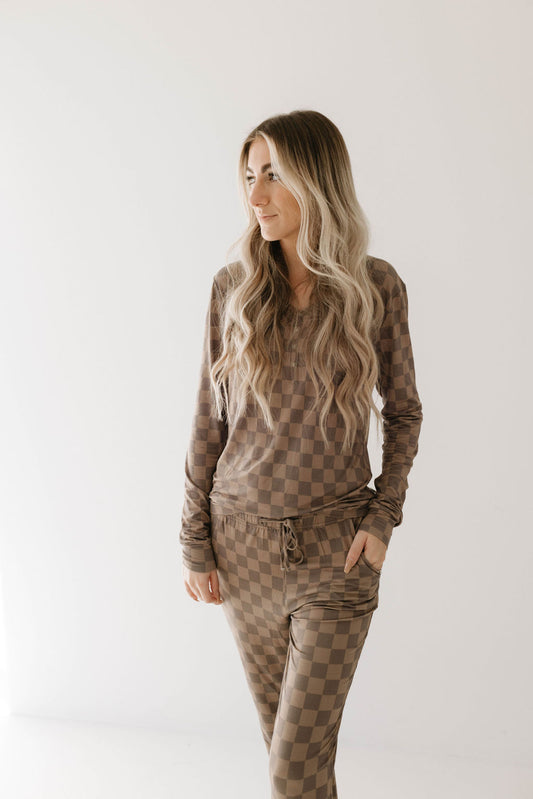 A woman with long, wavy blonde hair is standing and looking to her left. She is wearing the breastfeeding accessible Women's Bamboo Pajamas in Faded Brown Checkerboard by forever french baby, with her left hand in her pocket. The background is a plain white wall.