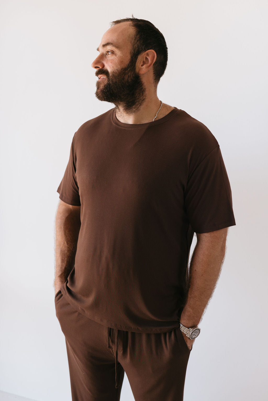 A man with a beard stands against a white background, looking to the side. He is wearing the Men's Bamboo Short Sleeve Pajamas in Coffee Bean by forever french baby, with his hands in his pockets.