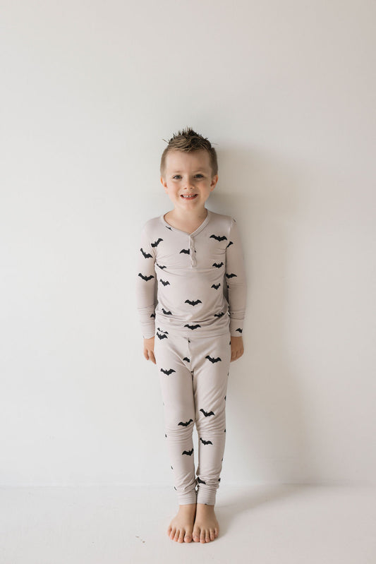 A young child stands barefoot against a plain white wall, smiling in their cream-colored "Bamboo Two Piece Pajamas | It's Bats!" from forever french baby. Their hair is short and styled with a messy, spiky look, radiating joy and comfort in the hypo-allergenic clothing adorned with a black bat pattern.