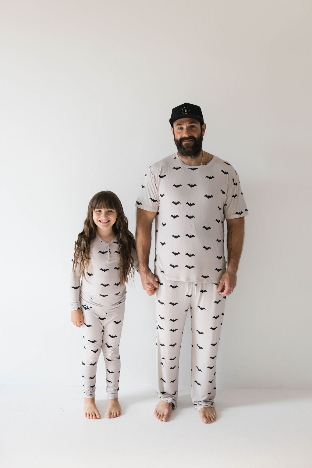 A bearded man wearing a black cap and a young girl with long hair and bangs stand holding hands against a plain white background. They are both dressed in matching hypo-allergenic Adult Bamboo Short Sleeve Pajamas from forever french baby, featuring ultra-soft bamboo fabric with a pattern of black bats on light-colored material.