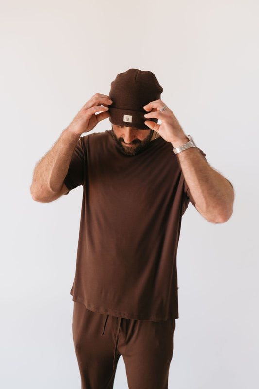 A person dressed in a matching ensemble of the Men's Bamboo Short Sleeve Pajamas in Coffee Bean from forever french baby adjusts their beanie. They are positioned against a plain white background.