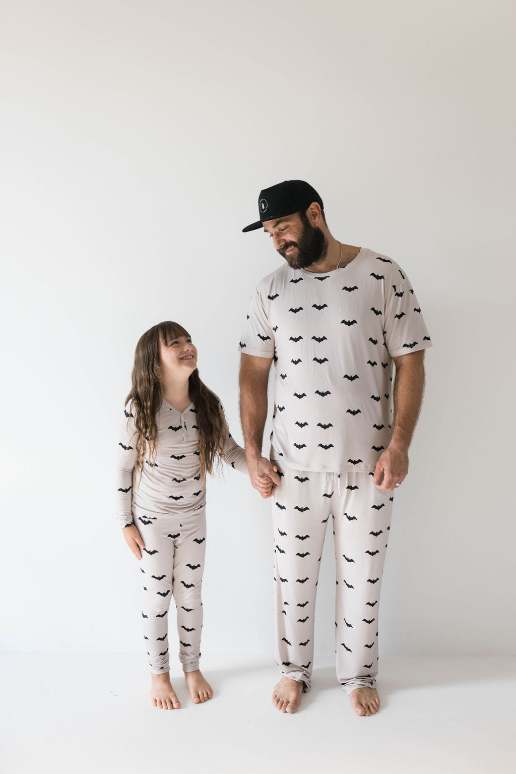 A man with a beard and a black cap and a long-haired child stand barefoot against a plain white background, smiling at each other. They are both wearing matching light-colored "Adult Bamboo Short Sleeve Pajamas | It's Bats!" by forever french baby. Holding hands, they enjoy the comfort of their ultra-soft, hypo-allergenic clothing adorned with bat prints.