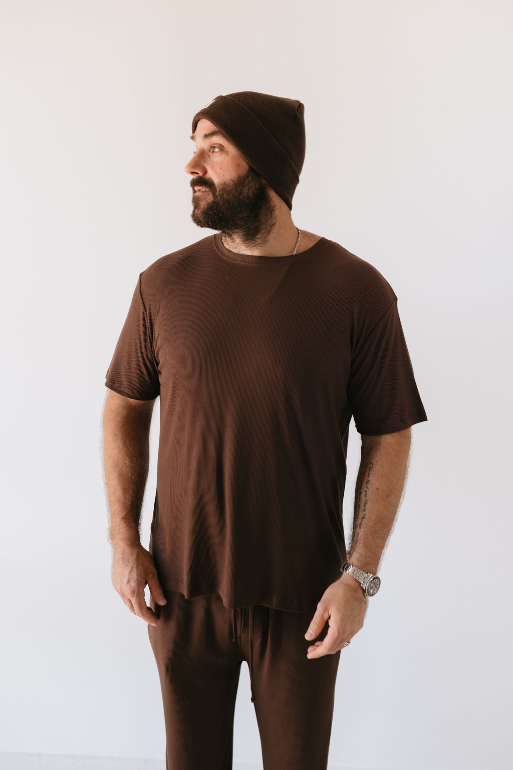 A man with a beard stands against a plain background, looking to the side. He is dressed in the Coffee Bean Men's Bamboo Short Sleeve Pajamas from forever french baby. His left arm features a tattoo, and he wears a watch.