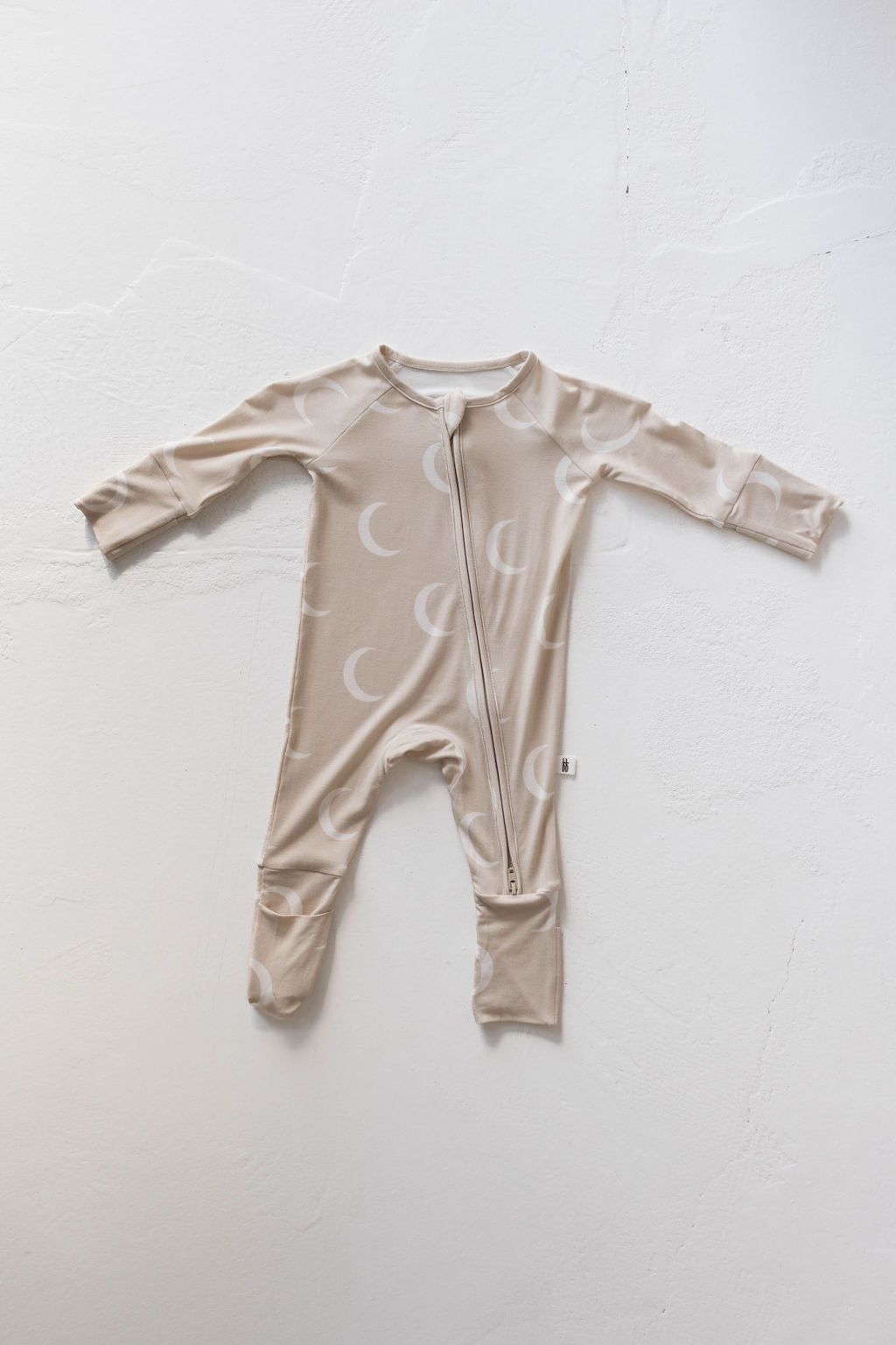 The Bamboo Zip Pajamas | Luna from forever french baby are the ideal baby sleepwear. This beige onesie features long sleeves and a front zipper, adorned with a charming white moon pattern. Made from hypo-allergenic fabric, it feels soft and comfortable against a light, textured surface.