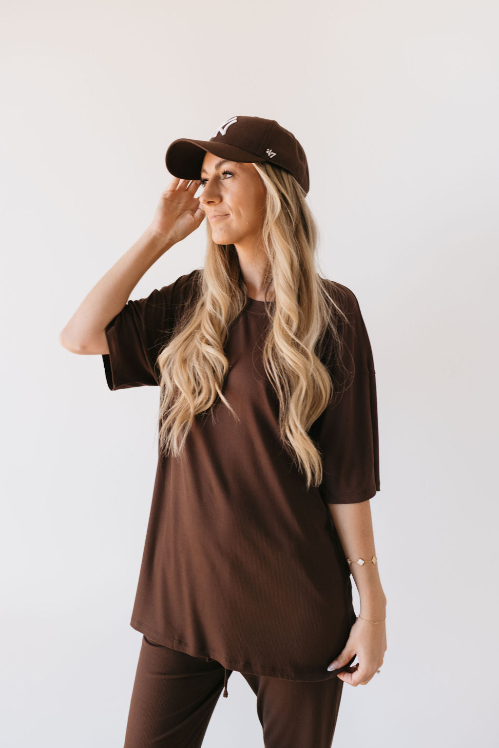 A woman with long blonde hair stands against a plain background, highlighting a comfortable ensemble. She wears the Short Sleeve Bamboo Women's Lounge Set in Coffee Bean by forever french baby, featuring a brown top and matching pants. She's looking to the side with a gentle smile.