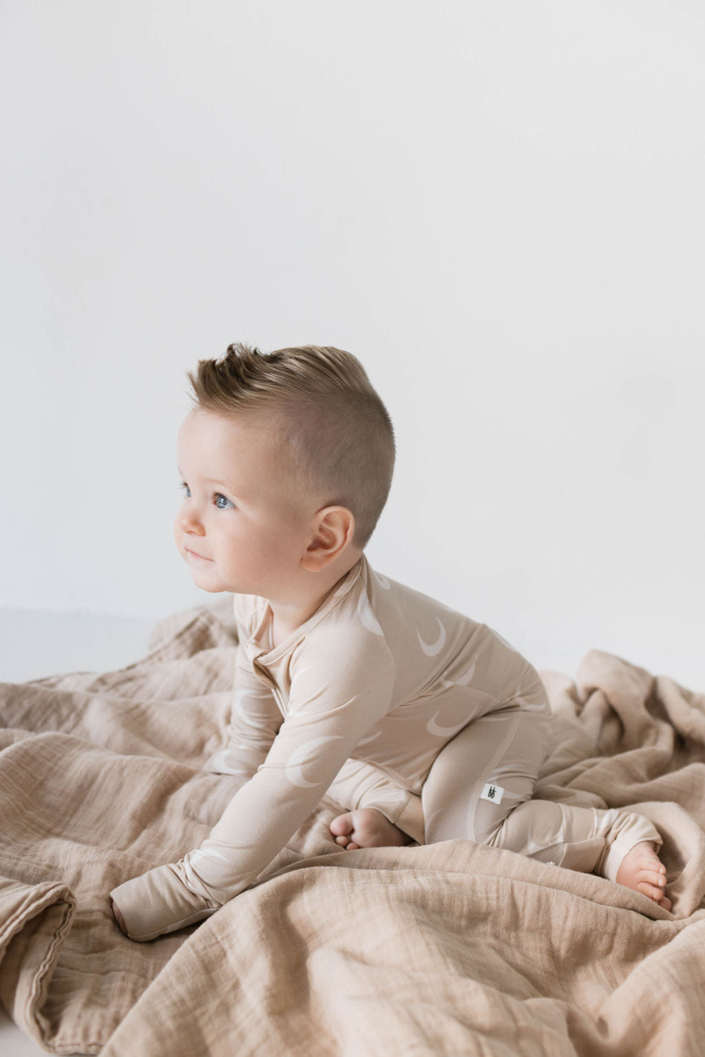 A light-skinned baby with short brown hair sits on a beige blanket, dressed in hypo-allergenic Bamboo Zip Pajamas featuring a charming moon pattern from forever french baby. The baby, looking to the left with a curious expression, is wearing the cream-colored Luna pajamas against a plain white background.