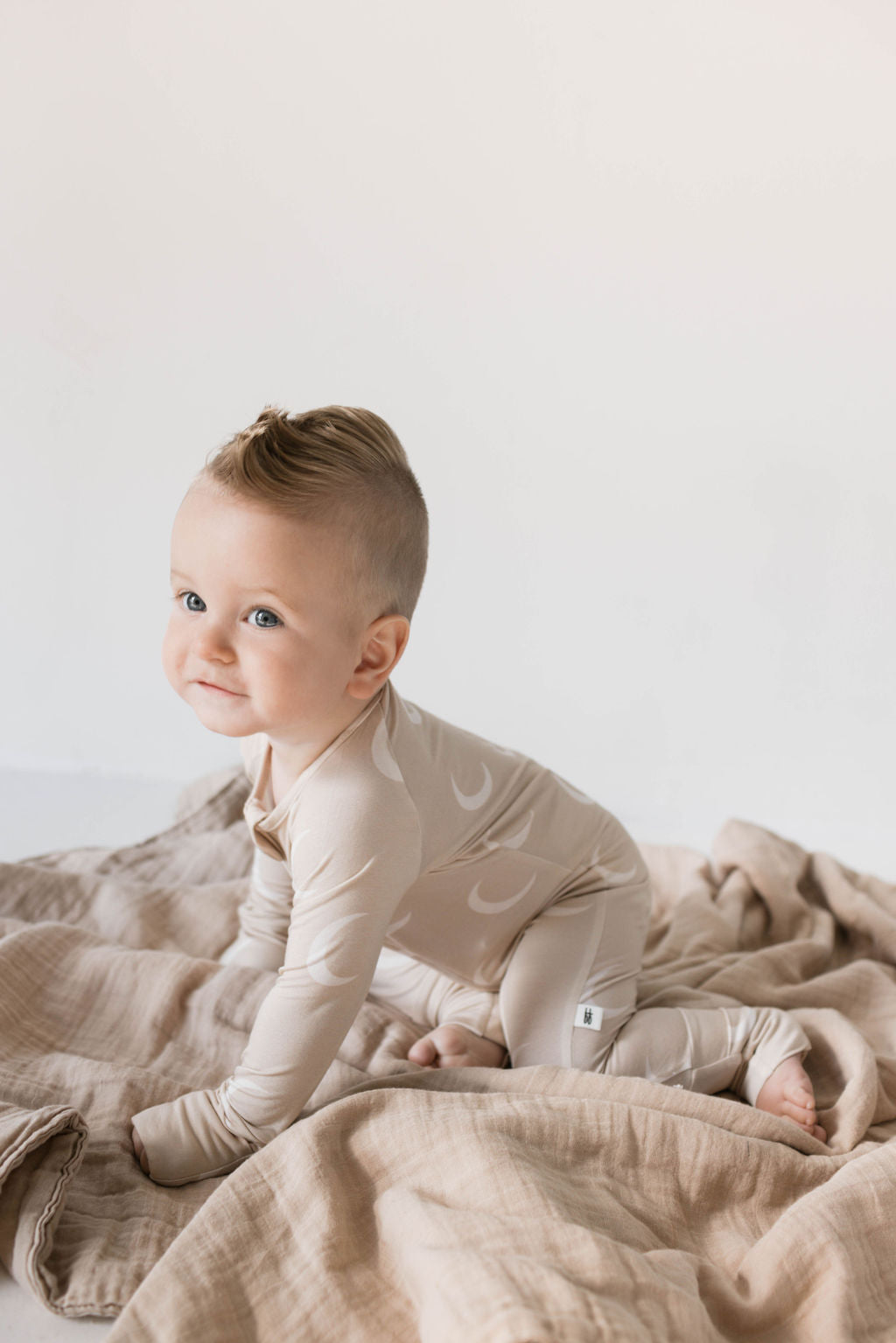 A light-skinned baby with light-colored hair is crawling on a soft, beige blanket. The baby is wearing forever french baby's hypo-allergenic Bamboo Zip Pajamas in the Luna design, featuring white crescent moons, and smiling while looking to the side. The background is plain and bright, drawing attention to the baby.