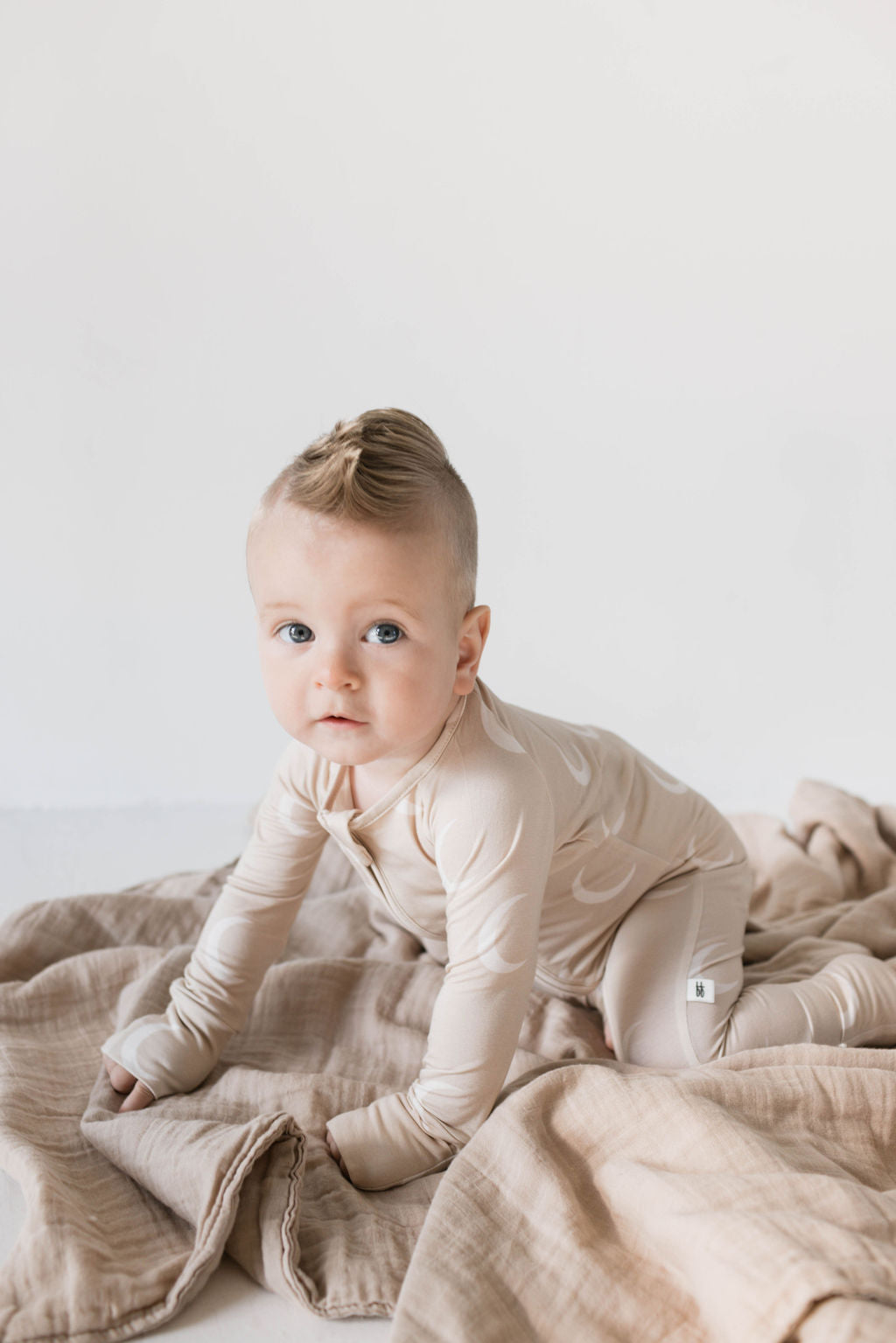 A baby with short hair and a small mohawk, dressed in hypo-allergenic **Bamboo Zip Pajamas | Luna** from **forever french baby**, is crawling on a beige blanket. The background is plain white.