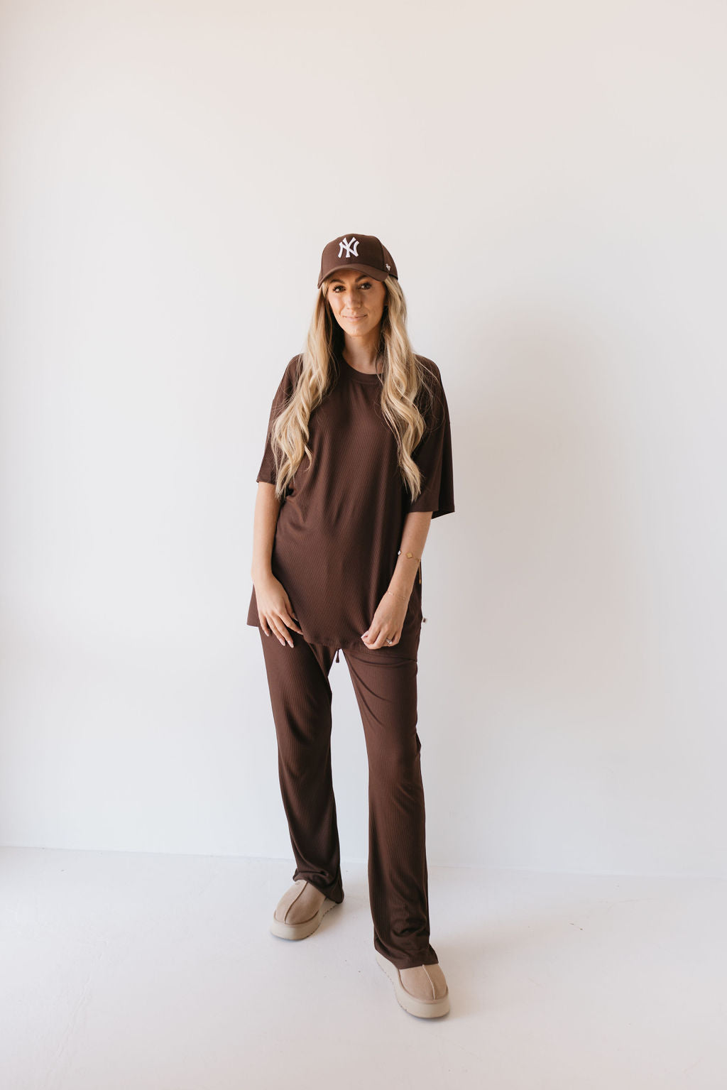 A person with long, wavy hair is wearing the "Short Sleeve Bamboo Women's Lounge Set" in the Coffee Bean color from "forever french baby." They stand against a plain white background, also sporting a brown baseball cap and tan sandals, enjoying the breathable fabric.