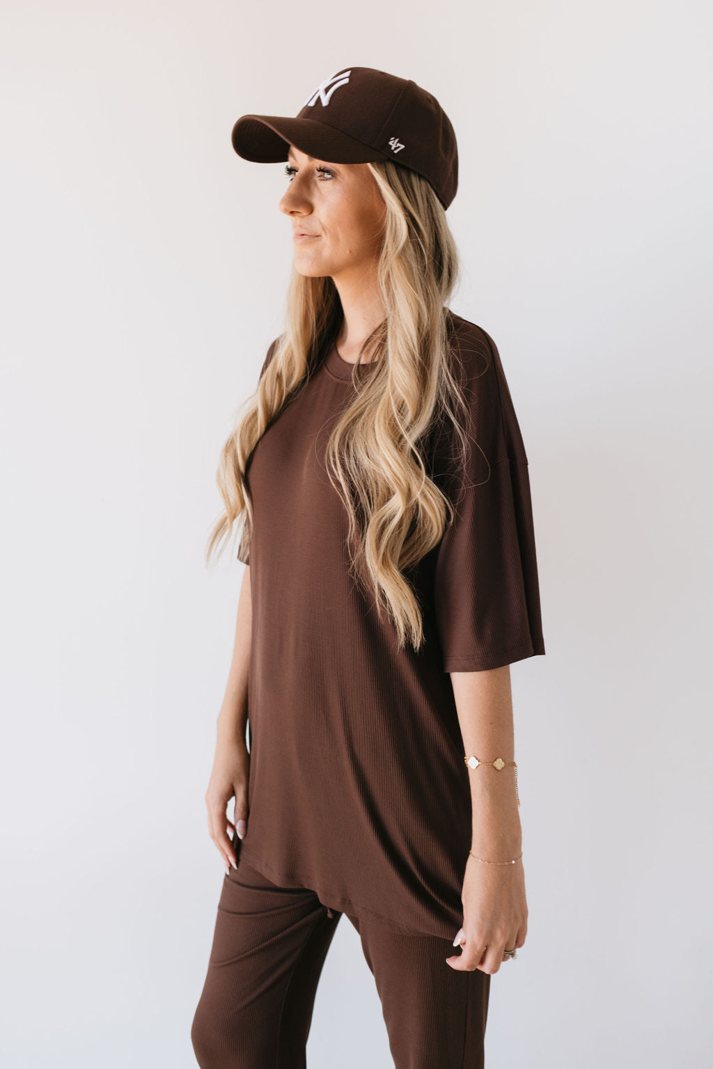 A woman with long blonde hair, wearing the Coffee Bean Short Sleeve Bamboo Women's Lounge Set by forever french baby and a brown cap, stands against a plain white background, looking to the side. Her left arm is adorned with a thin bracelet, adding elegance to her hypo-allergenic clothing ensemble.