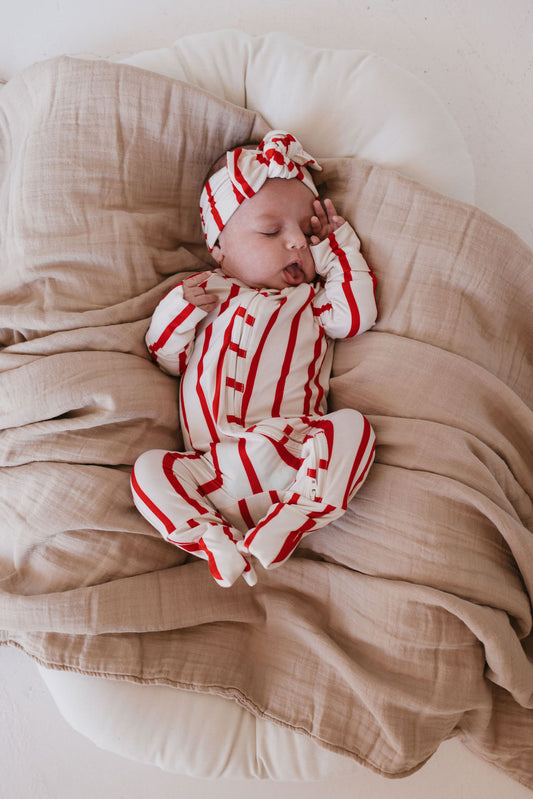 A baby sleeps peacefully on a beige cushion, wrapped in a soft, light brown blanket. Clad in the hypoallergenic Bamboo Zip Pajamas from forever french baby—a red and white striped outfit with a matching headband—the little one rests one hand gently on their head.