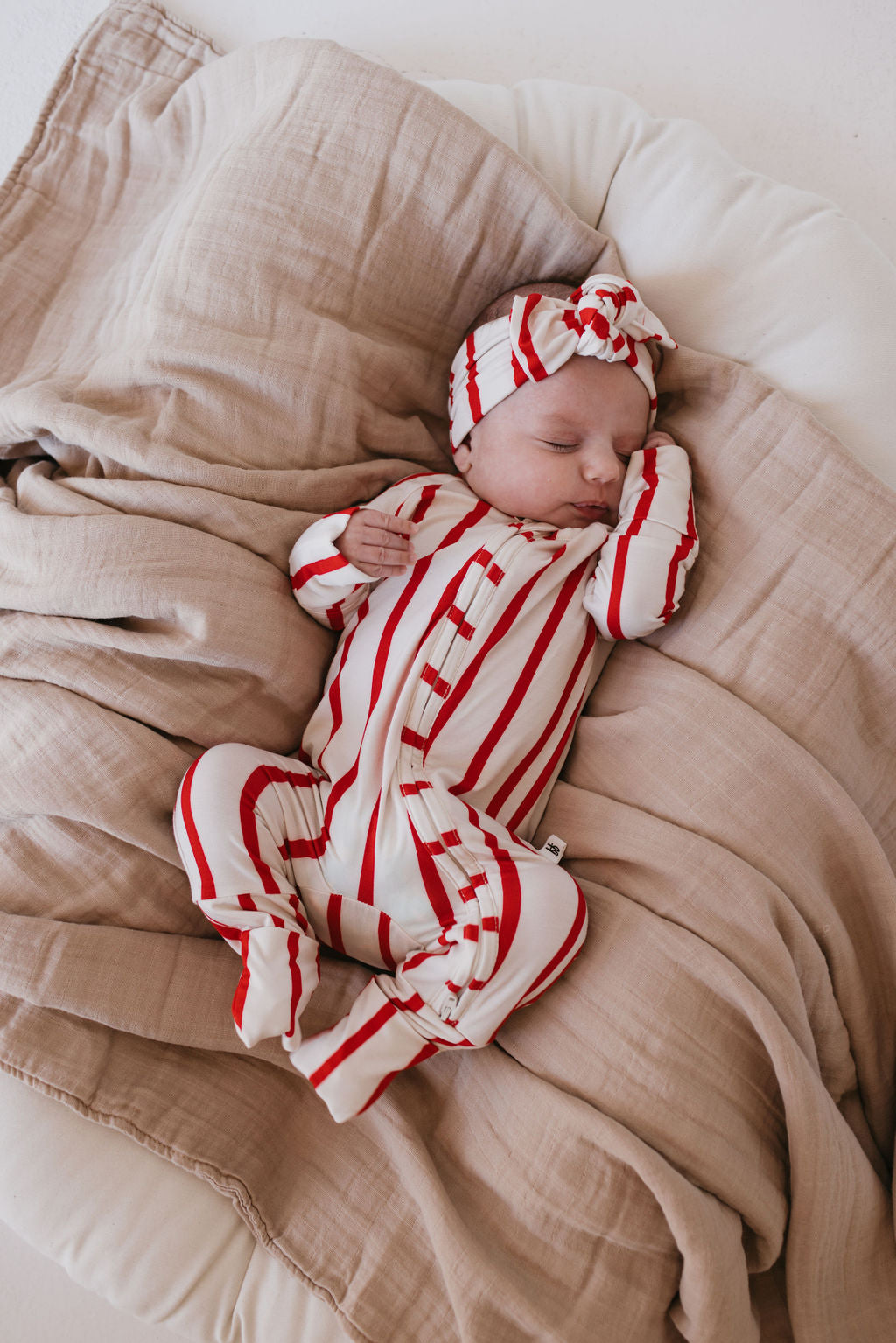 A baby sleeps peacefully on a beige blanket, wearing a red and white striped outfit with the matching Bamboo Head Wrap called "the Claus" by forever french baby.