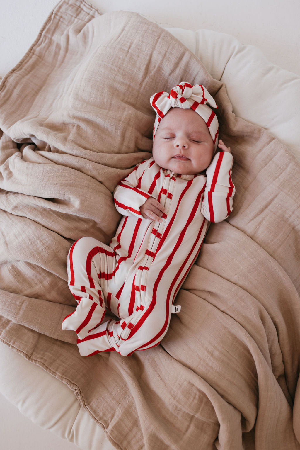 A sleeping baby in a red and white striped onesie and a cozy "the Claus" bamboo head wrap from forever french baby lies on a beige blanket.