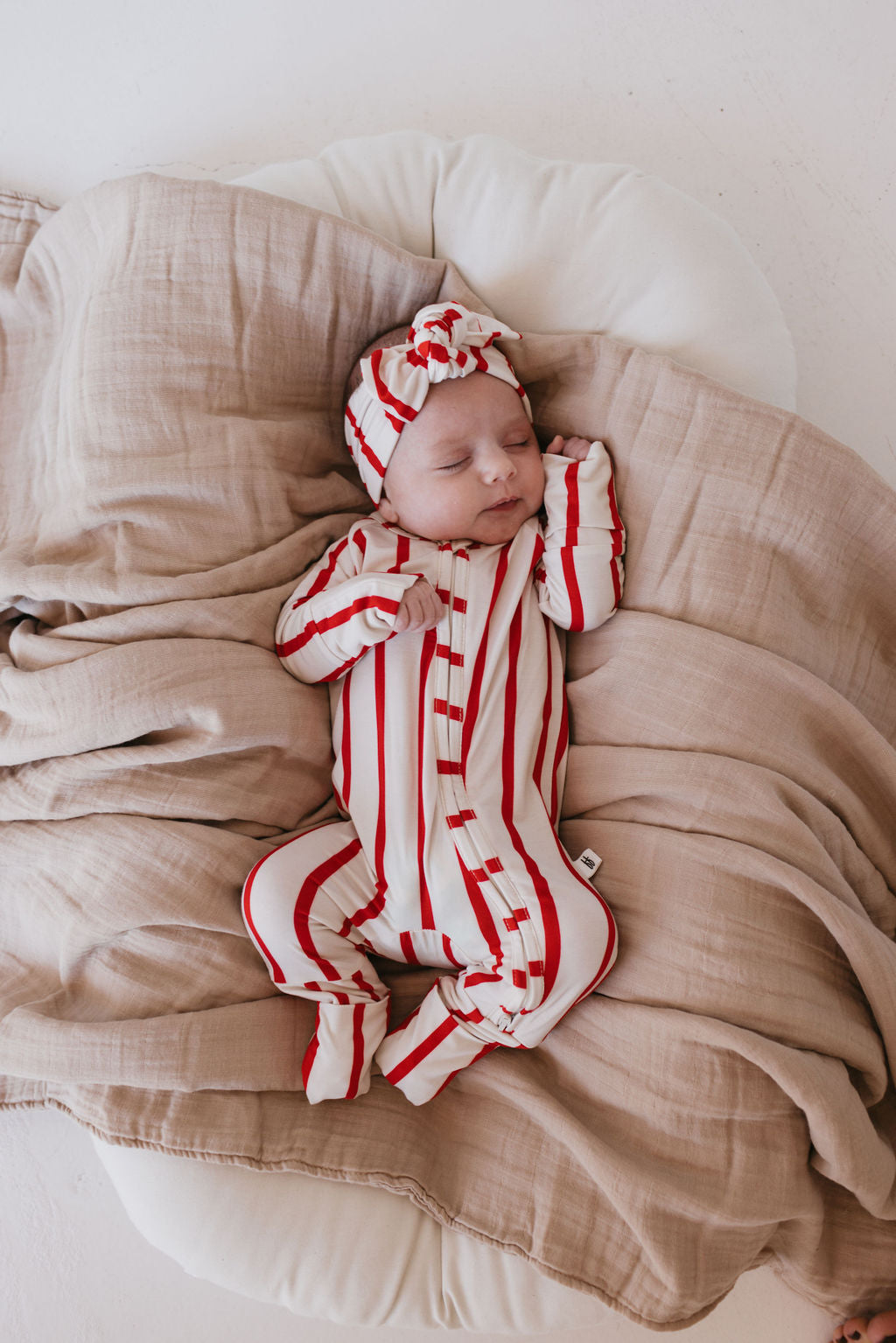 A baby sleeps peacefully on a soft, beige blanket, wearing a red and white striped onesie with an adjustable Bamboo Head Wrap called "the Claus" by forever french baby. Resting in a cushioned setting, this charming headband adds a touch of style to the serene scene.