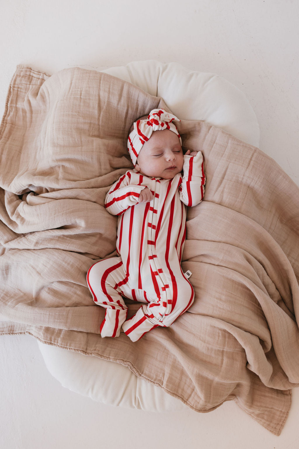 Snuggled under a soft, beige blanket, a baby sleeps peacefully, dressed in the red and white striped "the Claus" outfit, complemented by the matching Bamboo Head Wrap from forever french baby. Resting on a round cushion, the tranquil scene is made even cozier with carefully selected baby and toddler accessories.