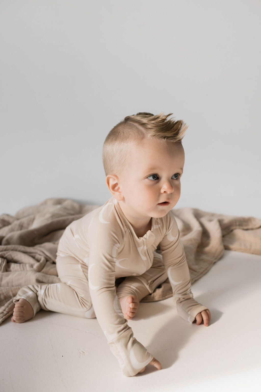 A baby with a short, stylish haircut and wearing full-length Beige Bamboo Zip Pajamas from Forever French Baby sits on a white surface, with a soft beige blanket in the background. The baby has an engaging expression and is looking slightly to the side.