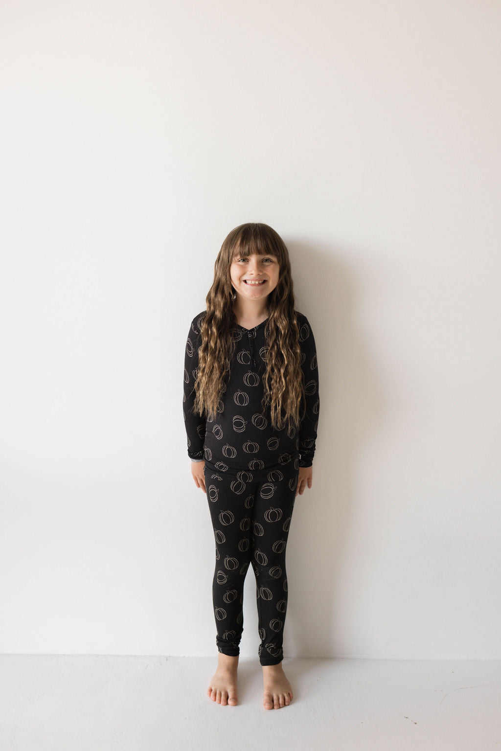 A young girl with long, wavy hair stands barefoot against a plain white wall. She is smiling and wearing the Bamboo Two Piece Pajamas | Midnight Pumpkin from forever french baby, showcasing breathable kids wear with a circular pattern.