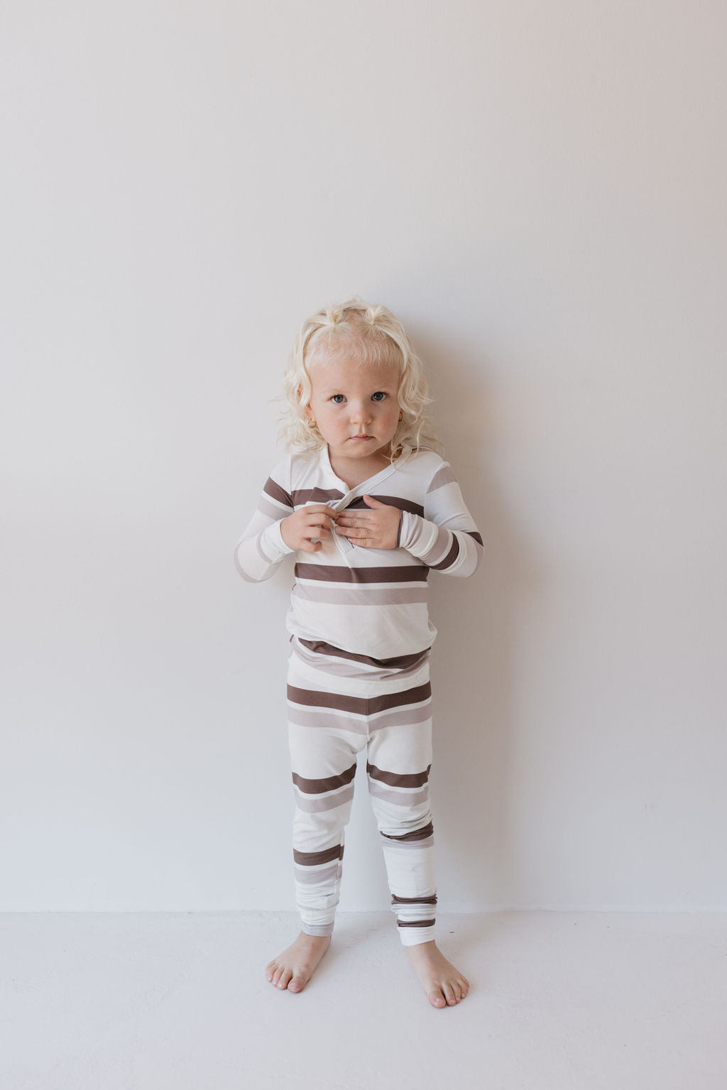 A young child with curly blonde hair stands barefoot against a white wall, dressed in the Bamboo Two Piece Pajamas | Woodland Stripes by forever french baby. The breathable fabric adds to their comfort as they adjust the top, a neutral expression on their face.