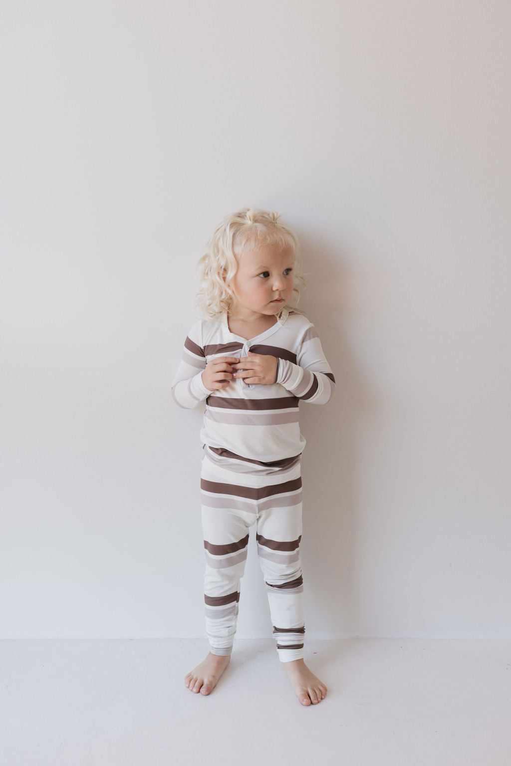 A child with curly blonde hair stands against a white wall, wearing Bamboo Two Piece Pajamas in Woodland Stripes by forever french baby. The striped outfit ensures both comfort and style as the child gazes to the side with hands clasped in front.