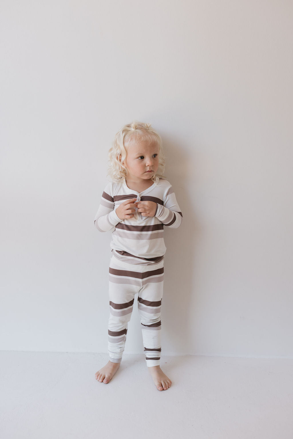 A young child with curly blond hair stands against a plain white wall, wearing the Bamboo Two Piece Pajamas in Woodland Stripes from forever french baby. With brown and white horizontal stripes, these hypo-allergenic pajamas are made from breathable fabric to ensure comfort. They look to the side with a neutral expression, their hands gently clasped.