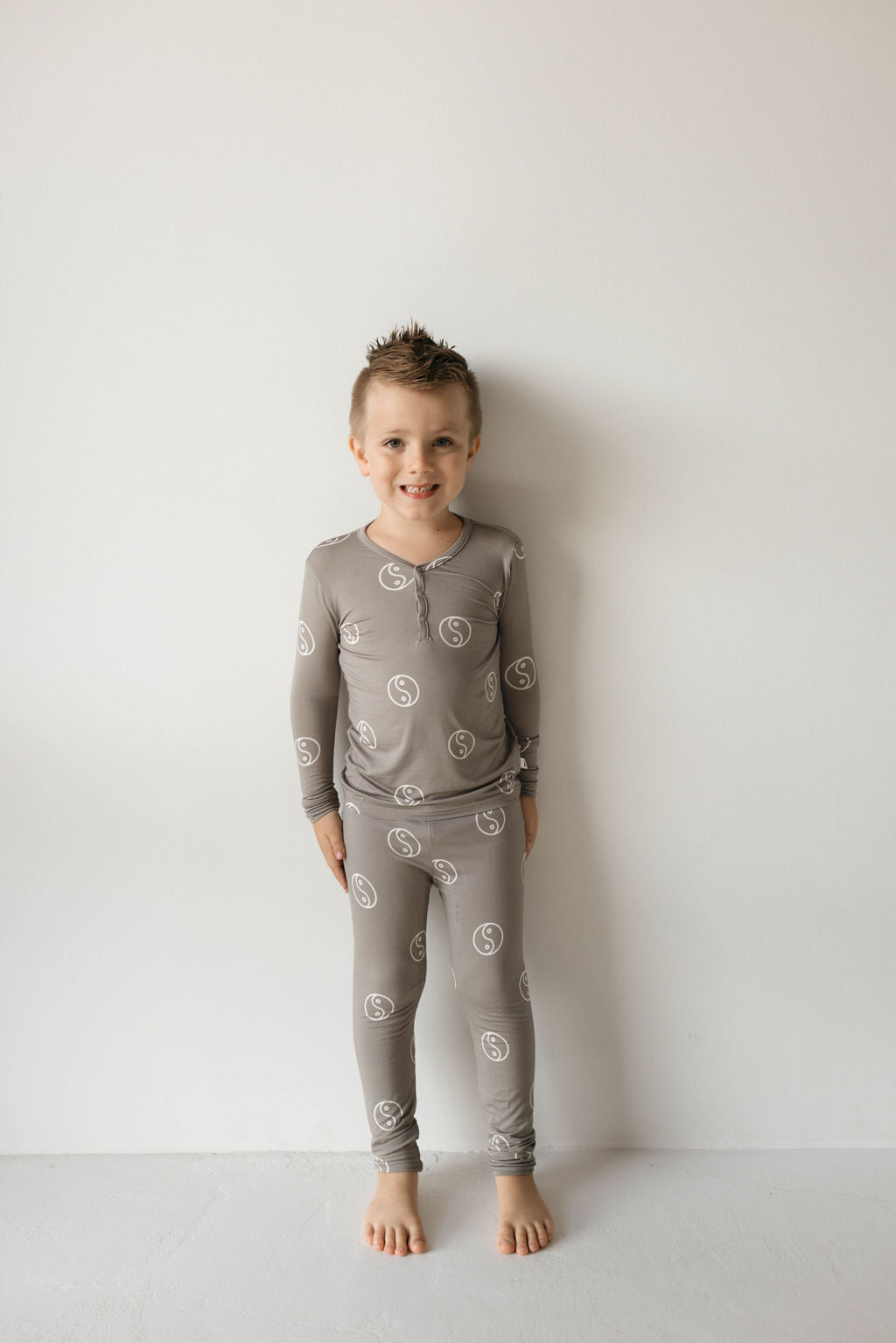 A young child stands against a plain white wall, smiling. The child is wearing matching Bamboo Two Piece Pajamas in Sage Yin Yang from forever french baby. Made from breathable bamboo fabric, the pajamas feature a repeating circular white pattern and include long sleeves and pants. The child is barefoot with short, spiked hair.