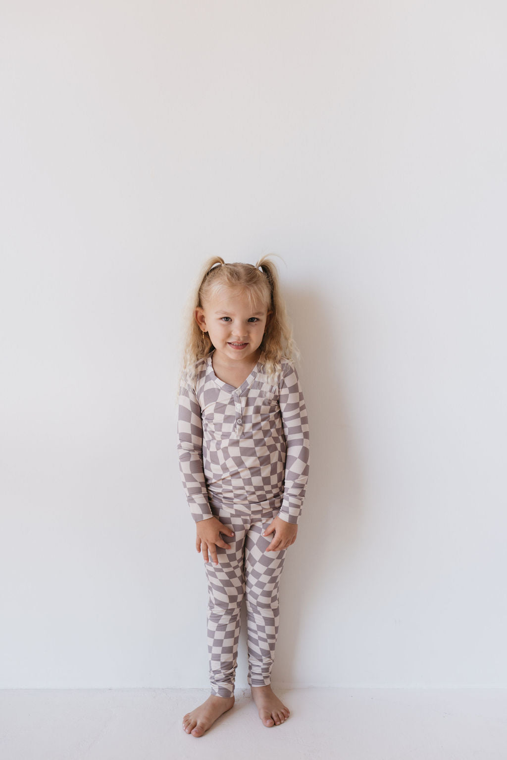 A little girl with light hair in pigtails smiles at the camera, standing barefoot against a plain white wall, while wearing hypo-allergenic Bamboo Two Piece Pajamas in Smokey Wave by forever french baby.