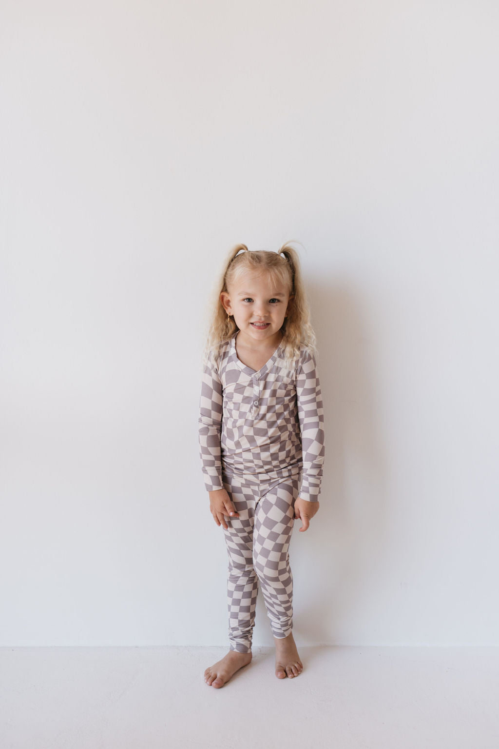 A young child with long blonde hair stands against a white wall, wearing the Bamboo Two Piece Pajamas in Smokey Wave from forever french baby. They are barefoot and smiling gently, with their hands resting at their sides.