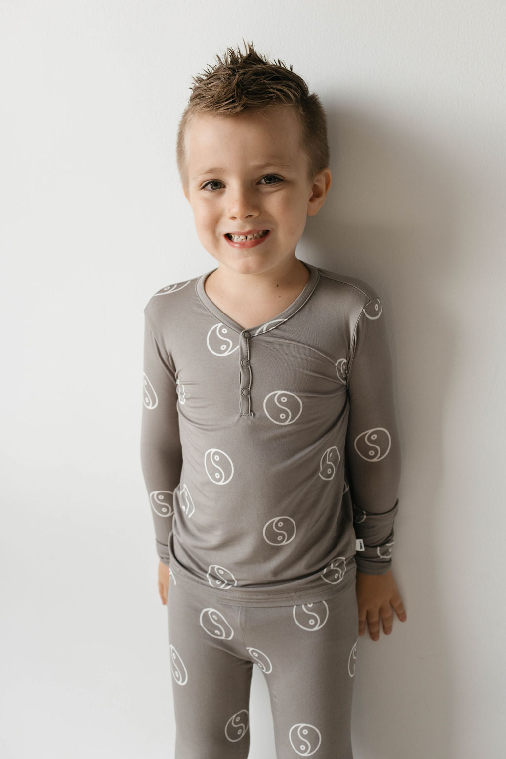 A young child with short spiky hair is smiling while standing against a white wall. The child is wearing the "Bamboo Two Piece Pajamas | Sage Yin Yang" by forever french baby, featuring matching gray pajamas made from breathable bamboo fabric and adorned with a repeating yin-yang symbol. The outfit includes a buttoned neckline, and the child poses with their hands by their sides.