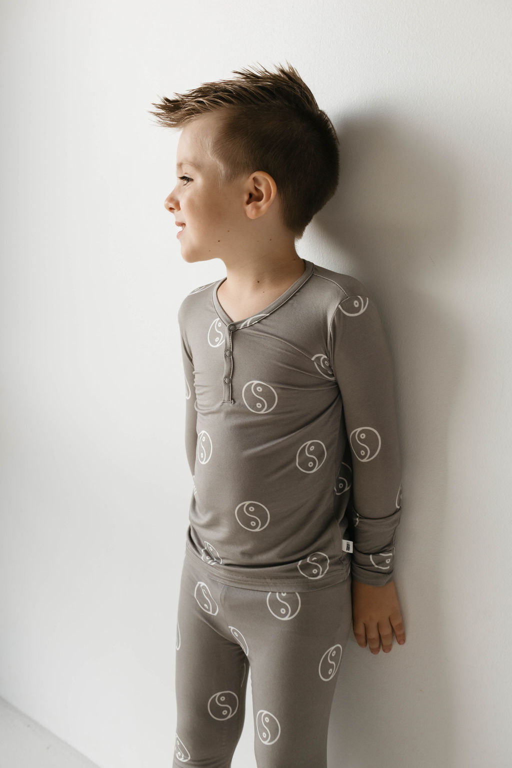 A young boy stands against a white wall, facing left and smiling. He is wearing the Sage Yin Yang Bamboo Two Piece Pajamas from forever french baby. His hair is styled in a spiky faux hawk.