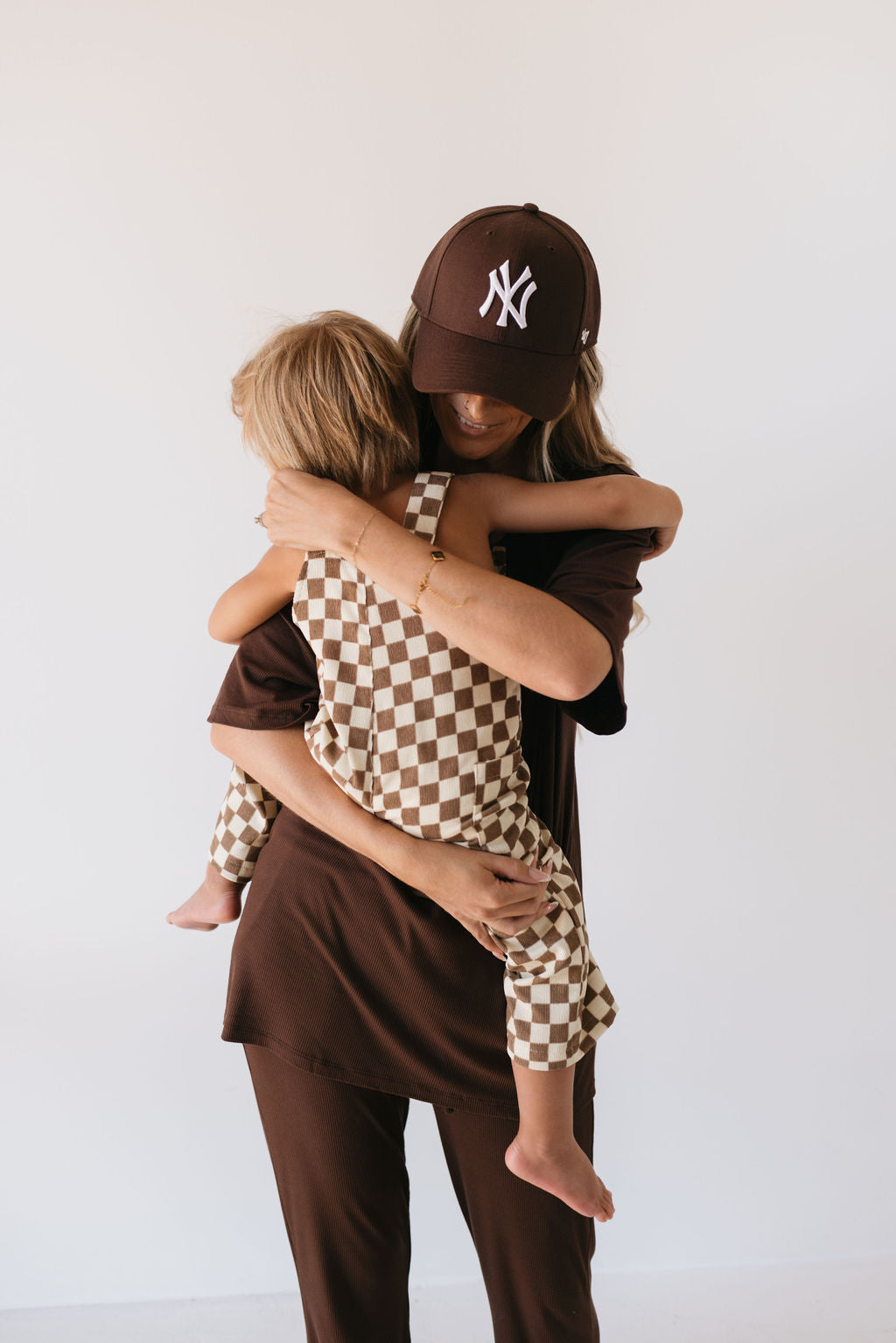 A woman wearing a brown baseball cap hugs a young child dressed in charming Corduroy Overalls featuring the Honeycomb Checkerboard pattern from forever french baby. They stand in a simple, neutral-colored setting.