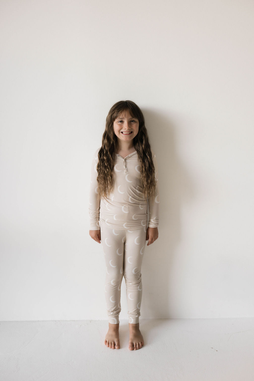 A young girl with long, wavy brown hair stands barefoot against a plain white background. She is smiling and wearing the matching light beige Bamboo Two Piece Pajamas | Luna by forever french baby, featuring a charming crescent moon pattern. These premium pajamas are celebrated for their breathable and hypo-allergenic properties.