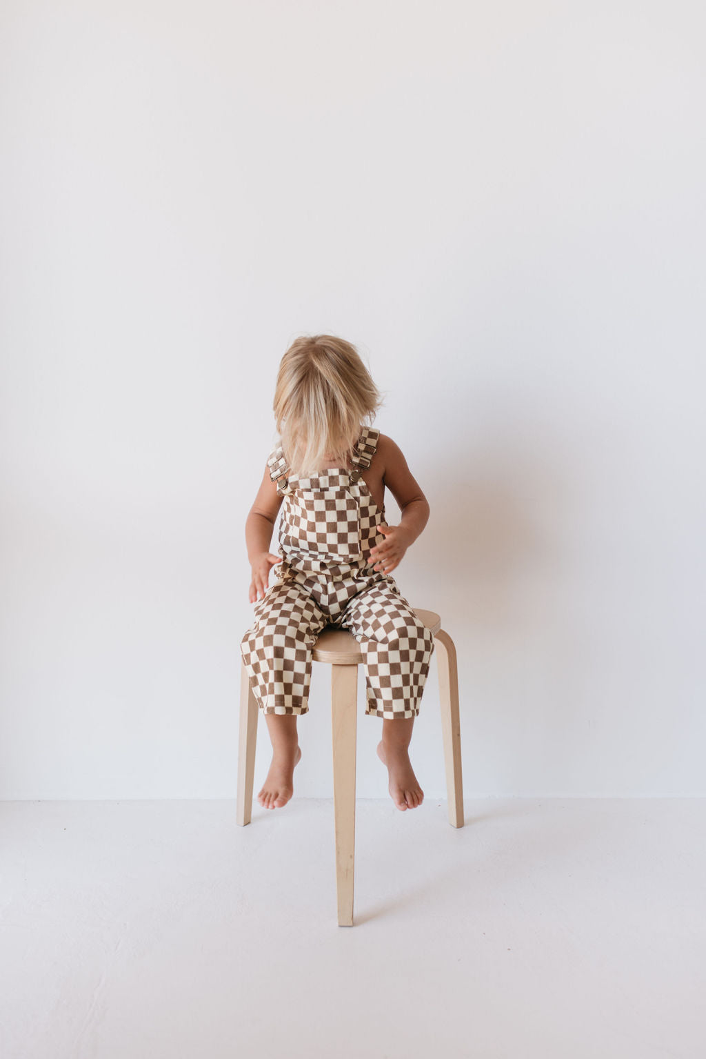 A child with blond hair is sitting on a wooden chair against a plain white background, wearing forever french baby's Corduroy Overalls in the Honeycomb Checkerboard pattern, and looking down at their hands.