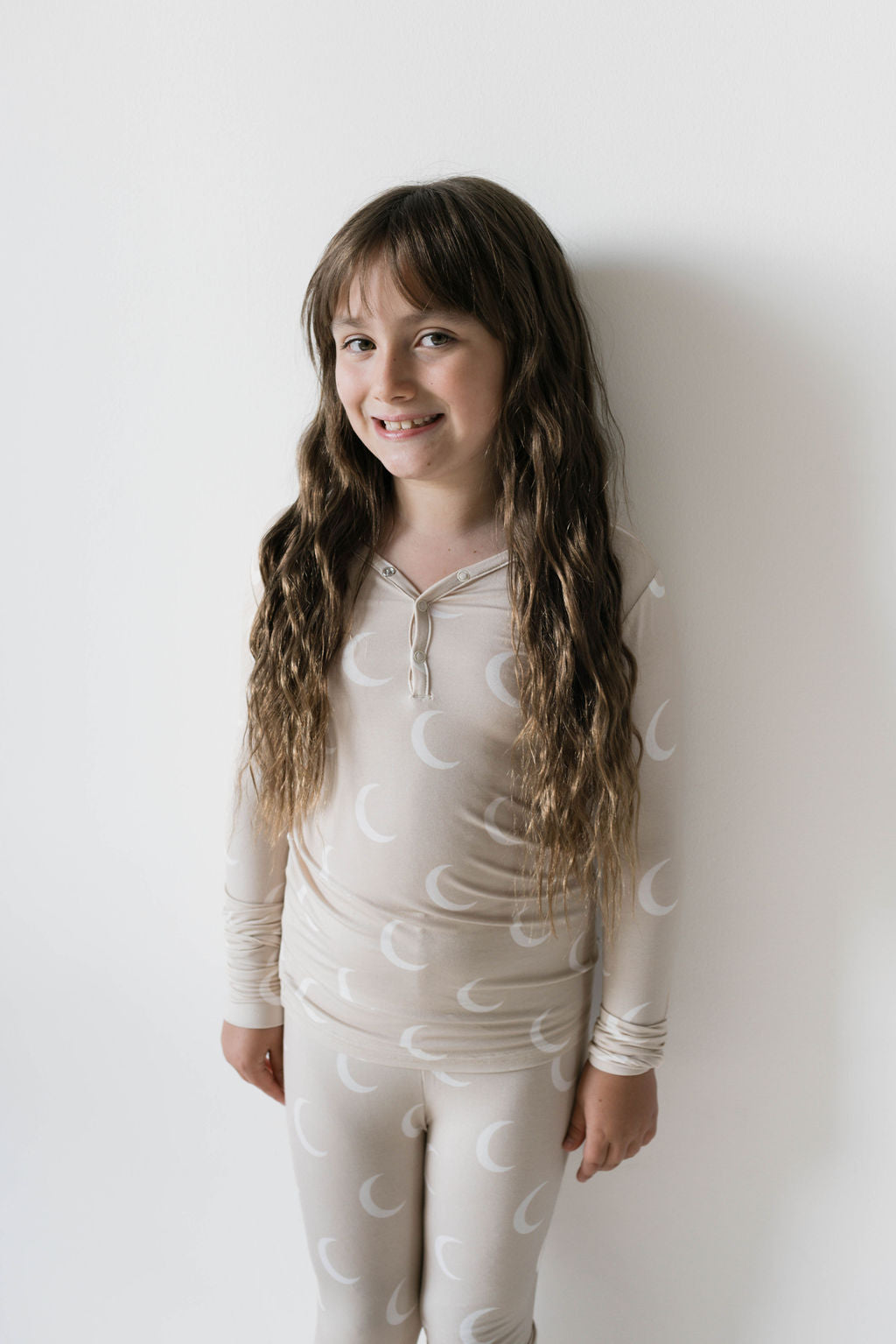 A young girl with long, wavy hair smiles at the camera. She is wearing bamboo two-piece pajamas called "Luna" by Forever French Baby, which feature a light beige outfit with a crescent moon pattern made from hypo-allergenic fabric. She stands against a plain white background.