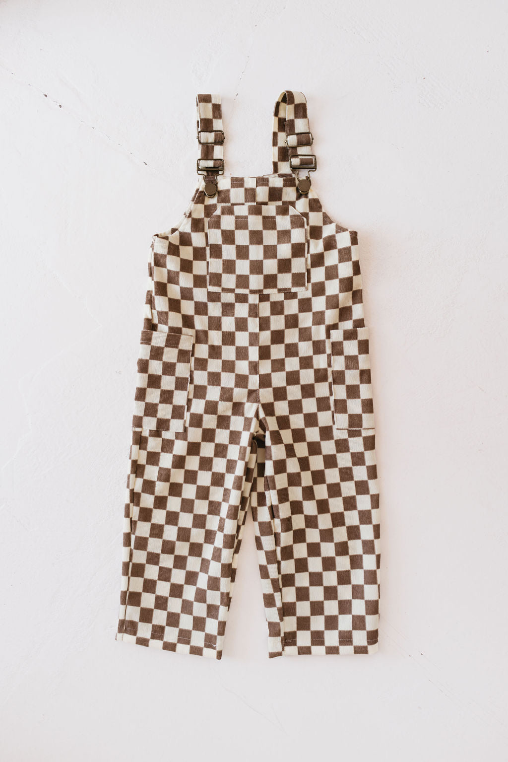 The Corduroy Overalls | Honeycomb Checkerboard by forever french baby are displayed flat on a white surface, showcasing straps fastened with metal buckles and large alternating squares in shades of brown and white. The soft corduroy fabric adds a textured feel to the pattern.