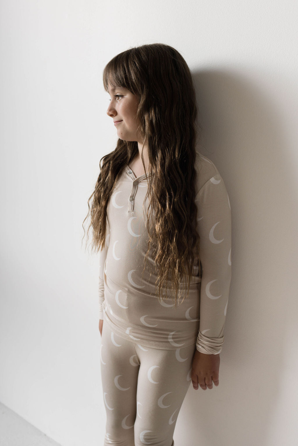 A young girl with long, wavy hair stands against a plain white wall, looking to the side. She is wearing light-colored, long-sleeved Bamboo Two Piece Pajamas | Luna by forever french baby, featuring a moon pattern.