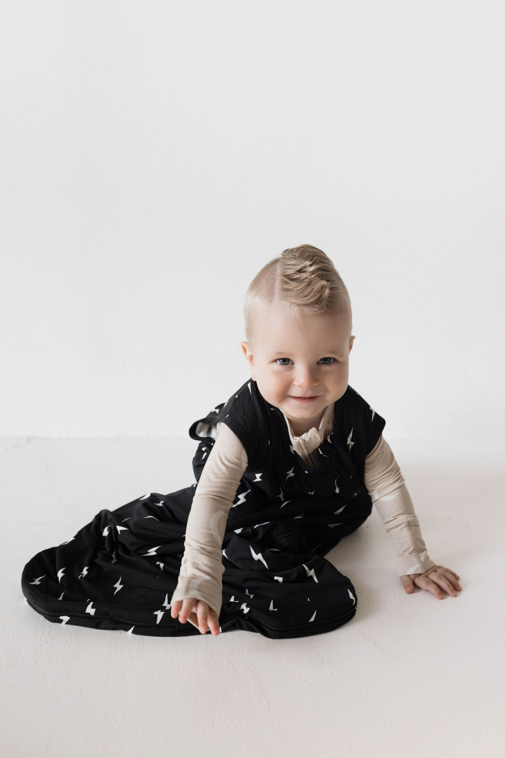 A baby with light hair sits on the floor, smiling at the camera. The baby is wearing the forever french baby's Bamboo Sleep Sack in Midnight Lightning Bolt over a cream-colored shirt. The background is plain and white, enhancing the luxurious sleep vibe.