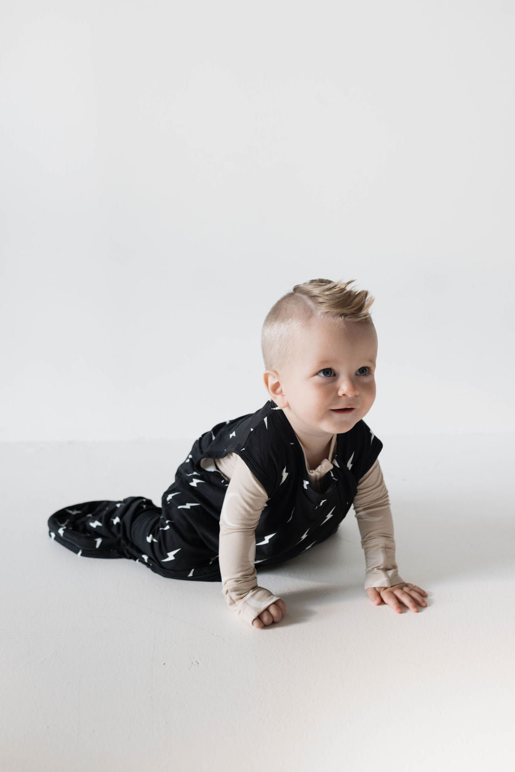 A baby with short blonde hair is crawling on a white surface. The baby is dressed in a black outfit with white bird-like patterns and a beige long-sleeve shirt underneath, wrapped snugly in the luxurious "Midnight Lightning Bolt" Bamboo Sleep Sack by forever french baby. The light background softly contrasts with the child's attire.