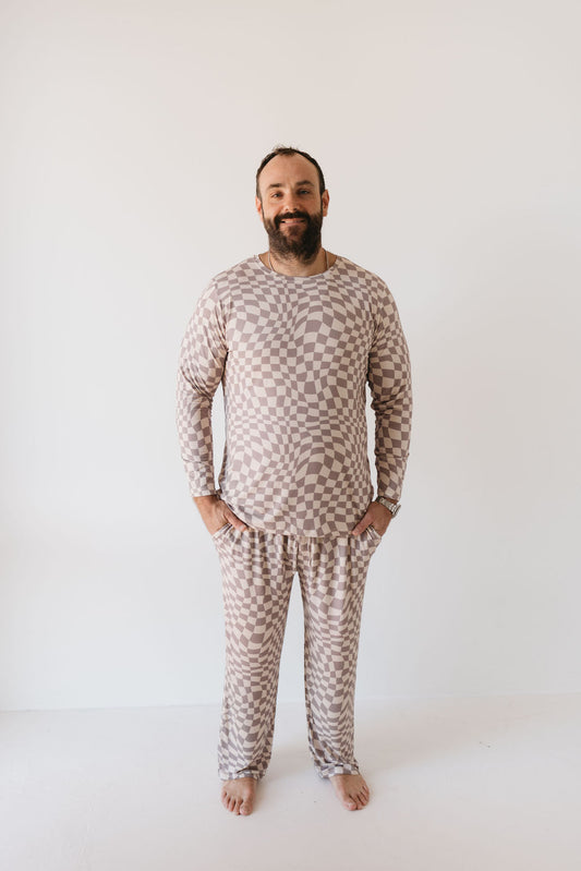 A man poses against a plain white background, dressed in the Smokey Wave Men's Bamboo Pajamas by forever french baby. The ensemble features a matching long-sleeve top and pants with a brown and beige checkered pattern. He is barefoot, with his hands in his pockets, smiling at the camera as he enjoys the comfort of the breathable fabric.