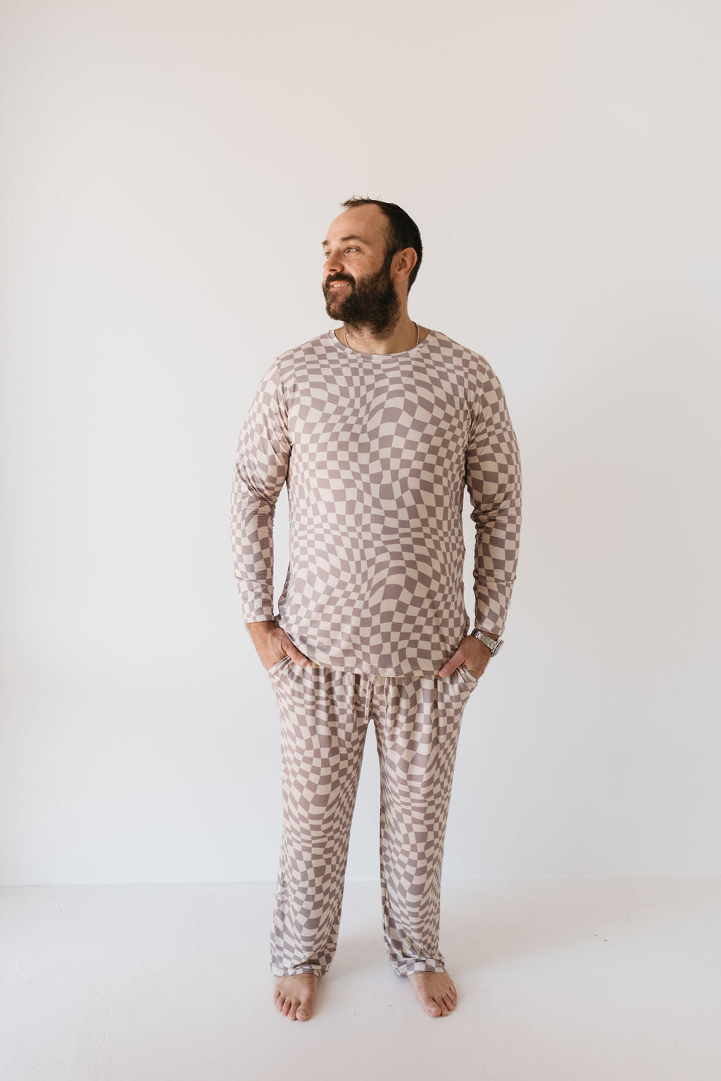 A man wearing the Smokey Wave Men's Bamboo Pajamas by Forever French Baby stands barefoot against a plain white background. With a relaxed expression and his hands in his pockets, he gazes off to the side, fully embracing the comfort of the breathable fabric.