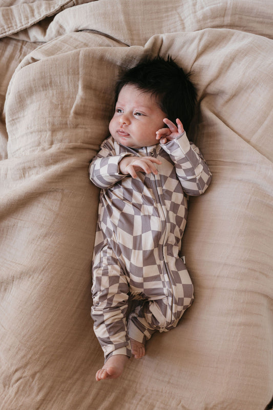 A baby wearing a Smokey Wave Bamboo Zip Pajamas from forever french baby lies on a soft beige blanket. The baby has dark hair and is looking to the side with one hand raised near the face.